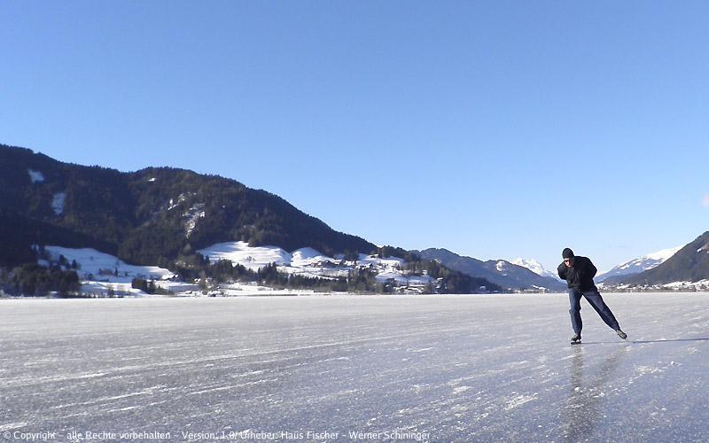Blick auf den zugefrorenen Weissensee mit den umliegenden Bergen und einem Schlittschuhläufer auf dem Eis.