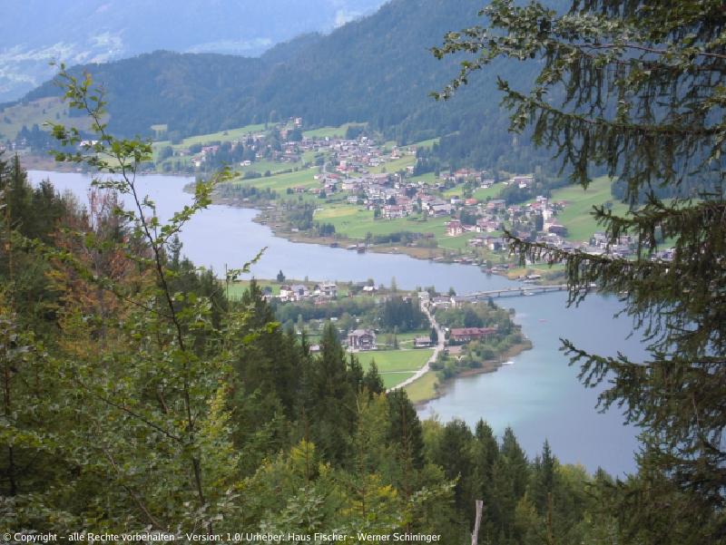 Blick auf den Weissensee, den Ort mit Brücke vom Wald aus zwischen Bäumen hindurch im Sommer.