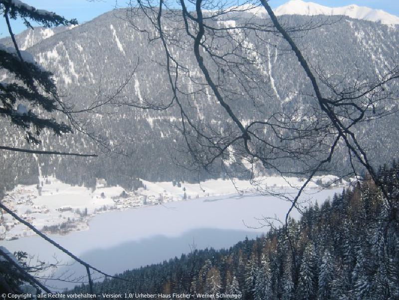 Blick auf den Weissensee im Winter aus einer höheren Position vom Wald aus mit einem Berg im Hintergrund.